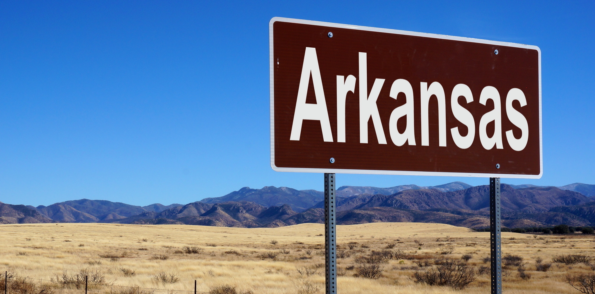 Highway sign showing the name Arkansas with mountains and field in the background.